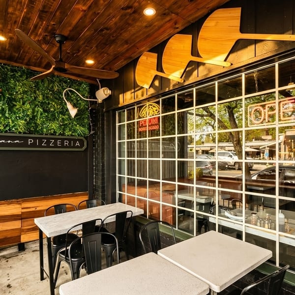 Outdoor patio of Romana Pizzeria with tables, chairs, a green wall, and a large window.