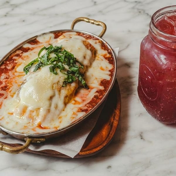 Baked pasta dish with melted cheese and basil, next to a red iced drink on a marble table.