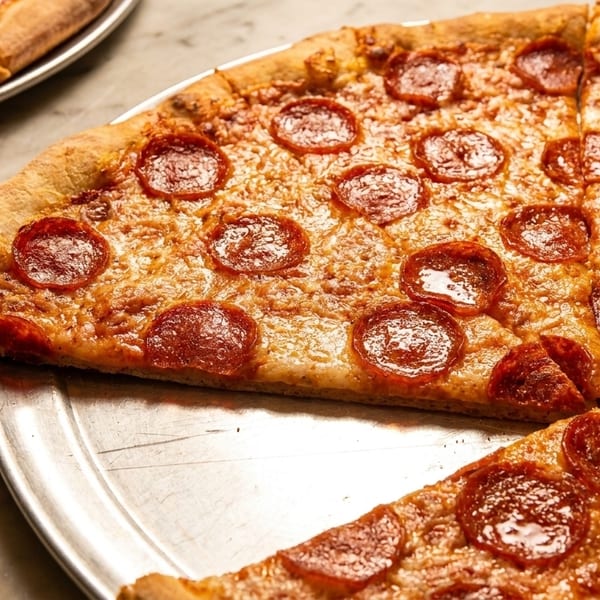 A freshly baked pepperoni pizza with a slice removed, on a metal tray, with another pizza in the background.